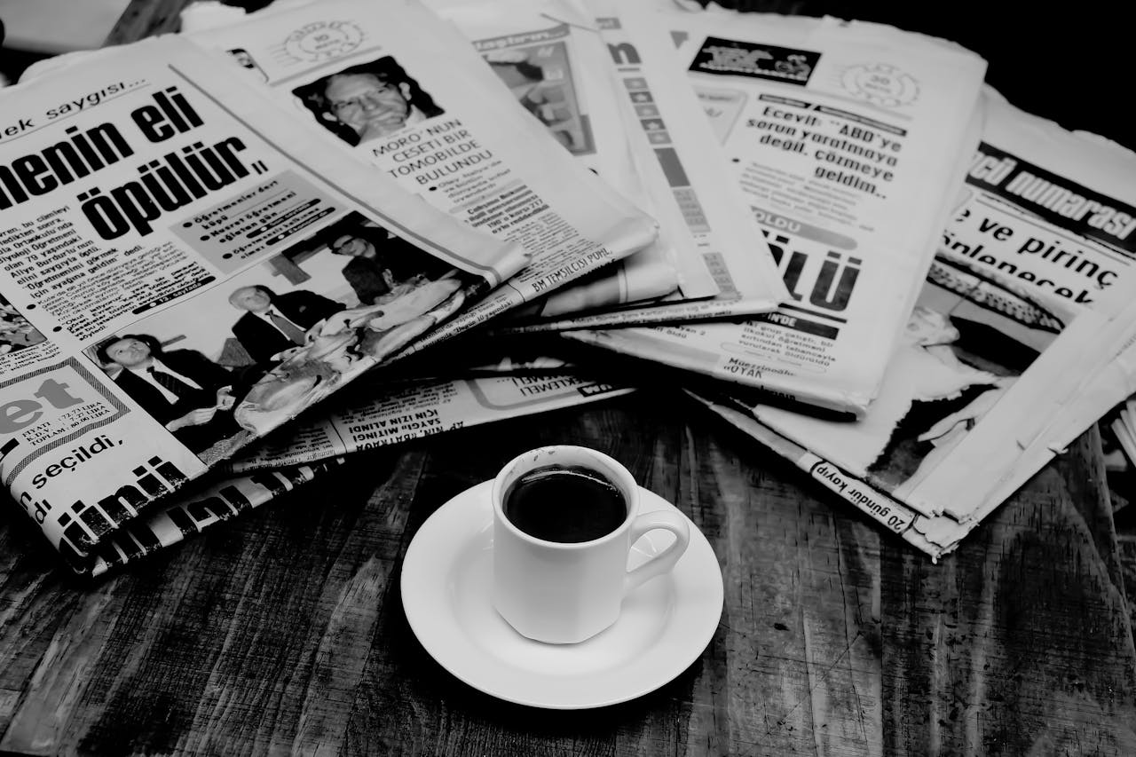 A cup of Turkish coffee sits on a wooden table next to a stack of newspapers.